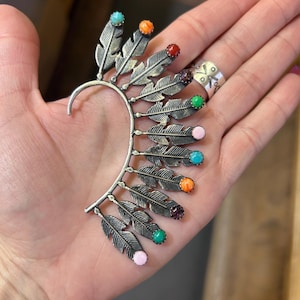 May include: A silver-toned ear cuff featuring a feather design. Each feather has a small, colorful gemstone at the tip, including turquoise, orange, red, purple, green, and pink. The cuff curves to fit the ear.