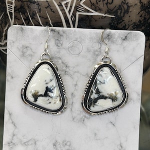 Può includere: Un paio di orecchini color argento con pendenti triangolari in pietra bianca. Le pietre presentano venature nere e grigie e sono incastonate in un bordo nero con un bordo argentato decorativo. Gli orecchini pendono da ganci d'argento.