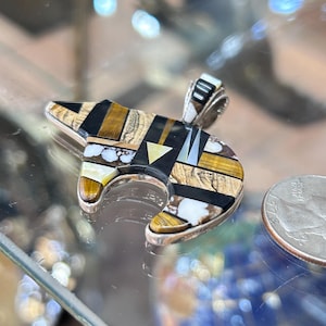 May include: A silver bear pendant with a mosaic design of black, brown, white, and yellow stones. The pendant is next to a coin. The pendant's design is intricate.