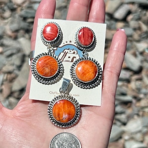 Puede incluir: Un conjunto de joyas con colgante, pendientes y anillo con detalles en naranja y plata. El conjunto incluye una moneda para comparar el tamaño. La joyería tiene un borde plateado en forma de cuerda alrededor de la piedra naranja.