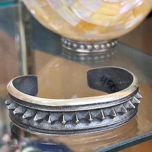 May include: A silver-toned metal cuff bracelet featuring a central row of pointed, triangular studs and a textured, darkened finish. The interior of the bracelet has the number 450 written in black ink on the inner surface of the metal band.