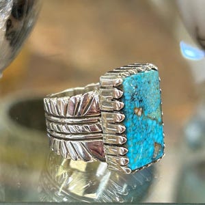 Puede incluir: Un anillo de plata con una piedra turquesa rectangular con patrones de matriz oscuros. La banda presenta un diseño texturizado de plumas repetitivas en los lados, que conduce a un engaste de plata dentado que asegura la piedra central.