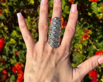 May include: A silver ring with a geometric design. The ring is wide and has a textured surface. The ring is worn on the middle finger of a hand.