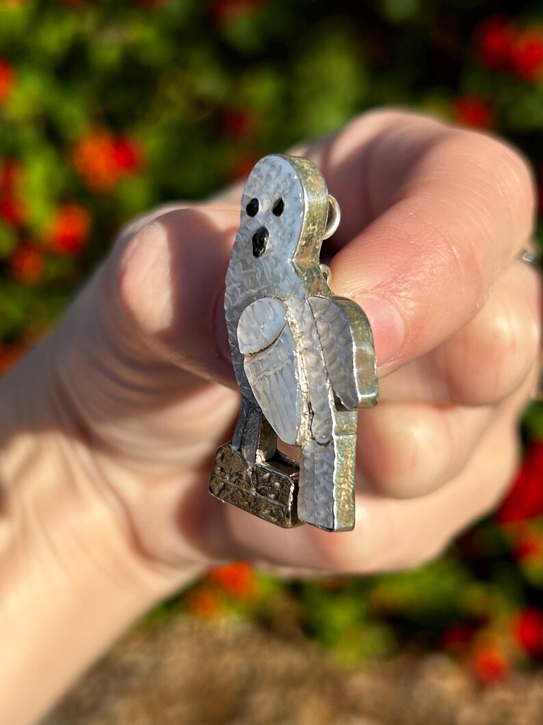 May include: A silver owl pendant with a mother of pearl inlay. The owl is perched on a branch with its wings spread out.
