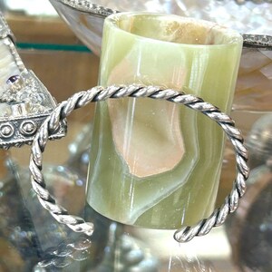 May include: A silver-toned cuff bracelet with a twisted rope design. The bracelet is open at the ends. The background includes a green onyx candle holder and a decorative silver-toned object.
