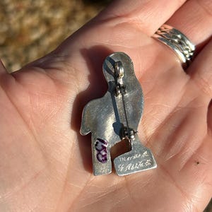 May include: Silver pin in the shape of a bird with a purple stone on the chest. The back of the pin has the inscription "Made in the U.S.A."