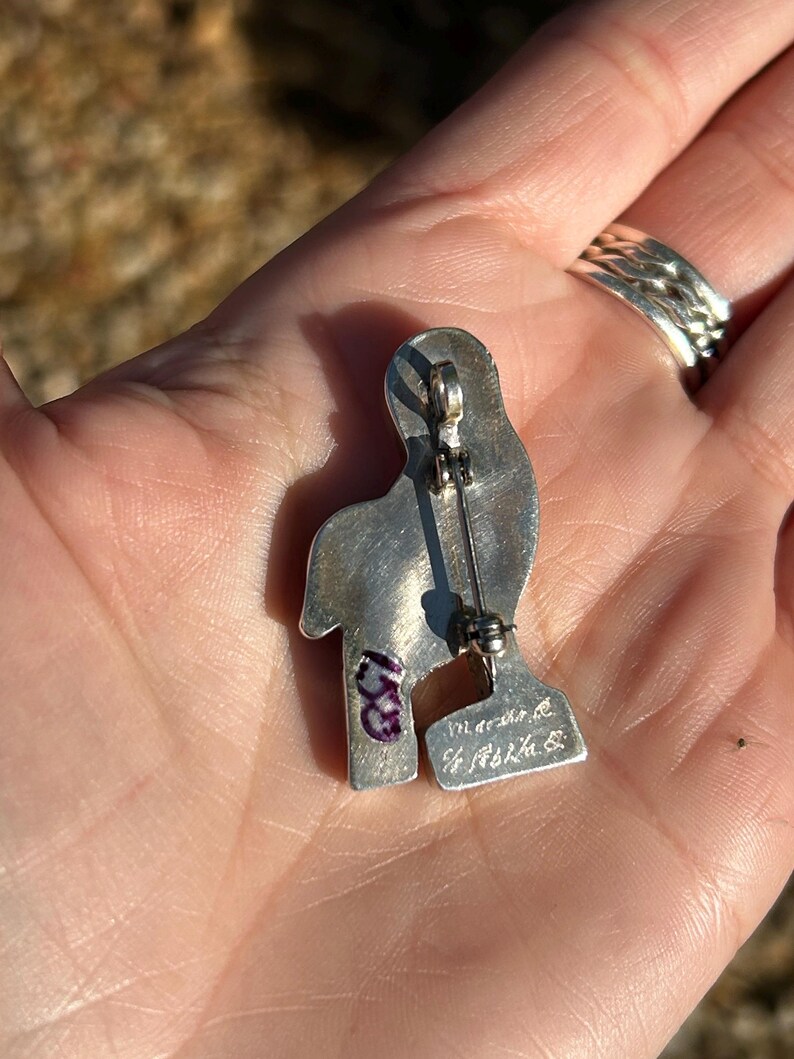 May include: Silver pin in the shape of a bird with a purple stone on the chest. The back of the pin has the inscription "Made in the U.S.A."