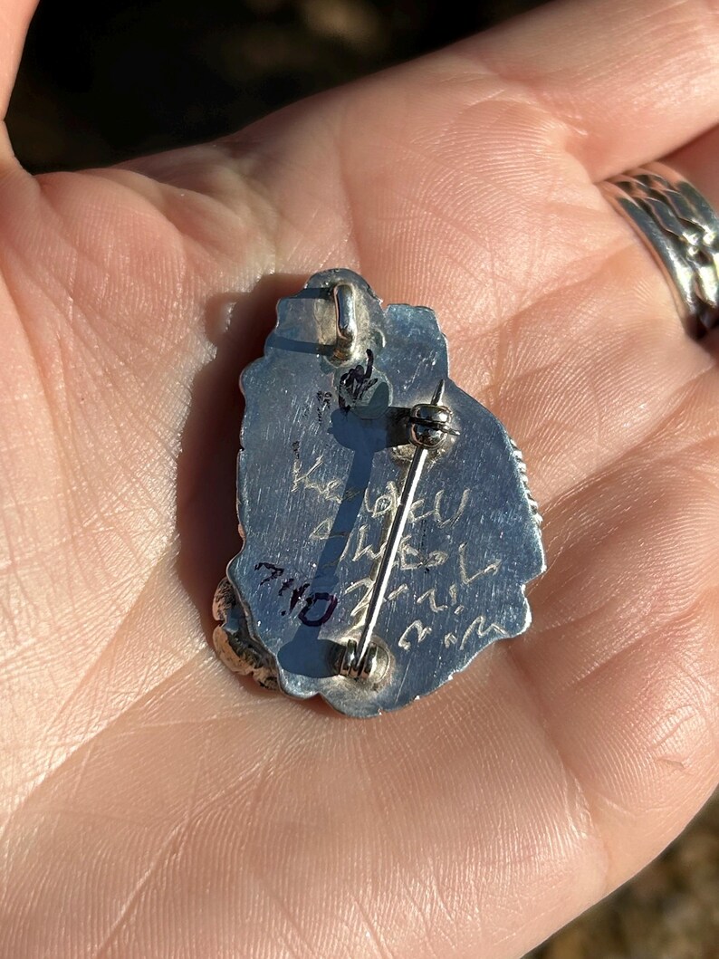 May include: Silver pendant with a detailed design and a pin back. The back of the pendant is inscribed with "7/10" and "Wesley".