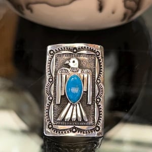 May include: A rectangular silver ring featuring a central turquoise stone inlay shaped like an oval. The silver surface is engraved with a stylised bird motif, surrounded by a decorative border with small circular and linear patterns.