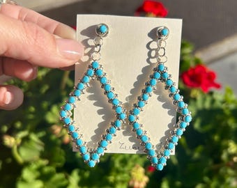 May include: A pair of silver dangle earrings with turquoise stones arranged in a diamond shape. The earrings are on a white card with the word "Zuni" written on it.