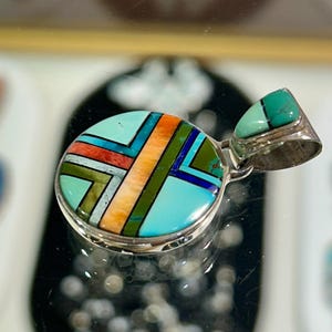 May include: A turquoise and silver pendant with a geometric design. The pendant features a round turquoise base with inlaid sections of orange, red, green, and blue. A smaller turquoise stone is set in the silver bail.