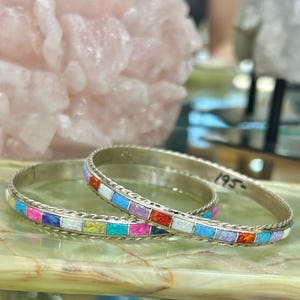 以下が含まれることがあります： カラフルな長方形の象嵌が施された2つのシルバーバングル。象嵌は、ピンク、ブルー、オレンジ、白など、さまざまな色を使用しています。バングルは、縁にねじれたロープのデザインが施されています。