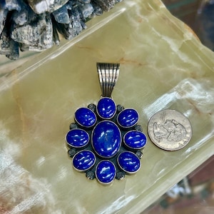 Puede incluir: Colgante de plata con una piedra ovalada central y piedras redondas azul oscuro circundantes. El colgante tiene una fianza decorativa de plata. Una moneda está al lado para comparar el tamaño. El diseño recuerda a una flor.