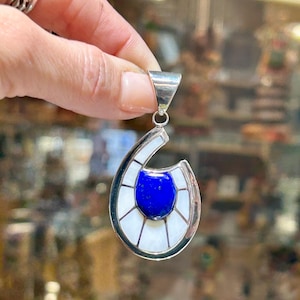 Puede incluir: Un colgante de plata con una piedra de lapislázuli azul rodeada de incrustaciones de nácar blanco. El colgante tiene forma de lágrima y tiene un aro de plata.