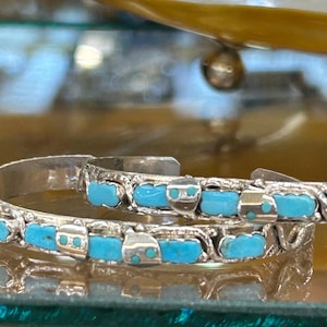 Puede incluir: Dos pulseras de puño plateadas con piedras de color turquesa. Las pulseras tienen un diseño calado decorativo. Las piedras turquesas son rectangulares y están engastadas en biseles individuales. Las pulseras se exhiben sobre una superficie reflectante.