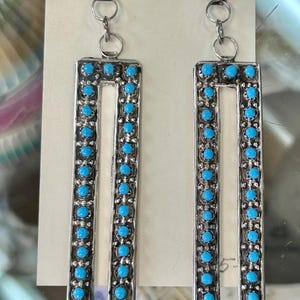 May include: A pair of silver-toned earrings with a rectangular design. Each earring features a row of small, round, turquoise-colored stones around the edges and a central white panel. The earrings have a small turquoise stone at the top and are connected by a chain.