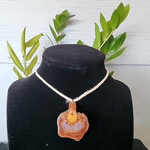 Puede incluir: Un colgante de cerámica marrón y blanco con forma de flor con un centro amarillo, colgado de un collar de cordón blanco sobre un expositor negro.