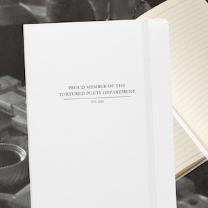 以下が含まれることがあります： 表紙に「Proud Member of the Tortured Poets Department Est 2024」と書かれた白いノート。ノートが開いており、罫線のあるページが見えます。