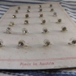 May include: A card of small, clear, rhinestone-studded buttons. Each button has a silver-coloured base and a domed, clear plastic cover. The card has a red border and the text "Made in Austria".