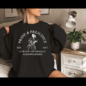 May include: Black sweatshirt with a white graphic print of a woman reading a book. The graphic includes the text "Pride & Prejudice", "Est. 1813", and "A Truth Universally Acknowledged".