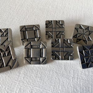 May include: Eight silver-toned metal buttons with geometric designs. Each button has a different pattern, including squares, triangles, and stars.