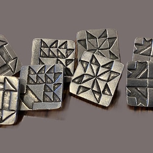 May include: A collection of seven square metal buttons featuring various embossed geometric patterns. Each button has a dark, brushed silver finish with recessed lines that create intricate shapes, including star and triangle motifs.