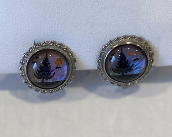 May include: A pair of silver-coloured stud earrings. Each earring has a round, domed design with a miniature landscape scene. The scene includes a silhouetted tree, a yellow sun, and a purple and blue sky. The edges of the earrings have a decorative, scalloped border.