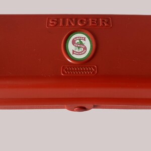 May include: A red Singer sewing machine case. The rectangular case has rounded edges and a clasp closure. The Singer logo is embossed on the top, along with a circular emblem and a decorative stitched pattern.