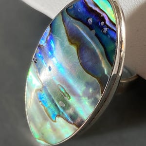 May include: An oval ring with an abalone shell displaying iridescent colours of blue, green, and purple. The shell is set in a silver-coloured metal band. The ring highlights the natural patterns of the abalone shell.