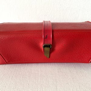 May include: A vintage red rectangular case with a textured surface. The case has a hinged lid with a pink strap and a brass-coloured clasp. The case is likely for storing small items.
