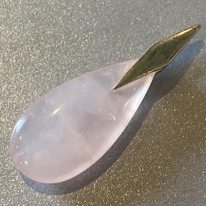 May include: A teardrop-shaped rose quartz pendant with a gold-colored metal accent. The pendant is a soft pink color and has a smooth, polished surface. The gold accent is a diamond shape and is attached to the top of the pendant.