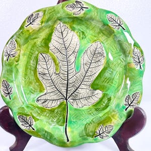 May include: A handcrafted ceramic plate with a fig leaf design. The plate features a vibrant green background with a large fig leaf in the center and smaller leaves around the rim. The plate is displayed on a wooden stand, with a real fig leaf in the foreground.