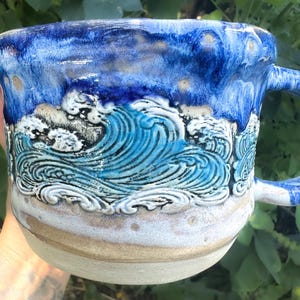 May include: A ceramic mug featuring a relief-carved ocean wave design. The upper rim is glazed in deep cobalt blue, transitioning to a textured, light blue and white wave pattern in the center, and a smooth, unglazed beige base at the bottom.