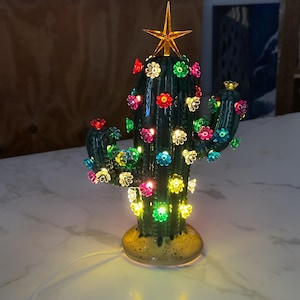 May include: A dark green ceramic cactus lamp featuring a glossy finish and a speckled tan base. The cactus is decorated with numerous small, multi-coloured translucent flower-shaped lights in pink, yellow, blue, and green, topped with a gold star.