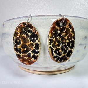 May include: A pair of ceramic earrings with a leopard print design. The earrings are oval shaped and have a silver hook.