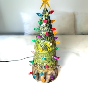 May include: A ceramic tabletop Christmas tree with a green glaze, featuring embossed sunflower and leaf patterns. The tree is decorated with small, multi-coloured plastic light bulbs and topped with a bright yellow, five-pointed star ornament.