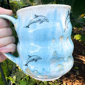 Może przedstawiać: Ręcznie robiony ceramiczny kubek z jasnoniebieskim szkliwem i teksturowaną powierzchnią. Kubek posiada czarne rysunki linii pływających delfinów. Uchwyt jest w pasującym jasnoniebieskim kolorze. Kubek jest trzymany na tle zielonych liści.