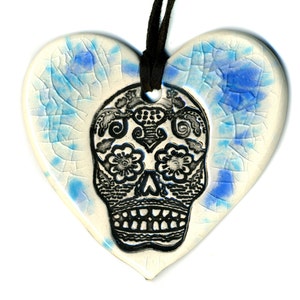 Op de afbeelding: Een hartvormig keramisch ornament met een gebarsten witte glazuurlaag en blauwe accenten. Een zwarte inktillustratie van een versierde schedel met bloem- en geometrische patronen staat in het midden. Een zwart koord zit door het bovenste gat.