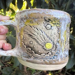 May include: A ceramic mug with a textured, beige-toned surface featuring a relief carving of a comet, stars, and a planet. The glaze includes accents of yellow and white, with a glossy finish on the rim and handle, showcasing a celestial design.
