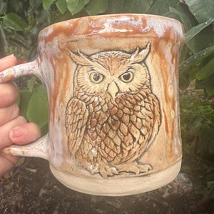May include: A ceramic mug featuring a raised, detailed relief of an owl on the front. The mug has a tan base color with a glossy, drippy brown and white glaze finish that cascades from the rim down the sides of the vessel.