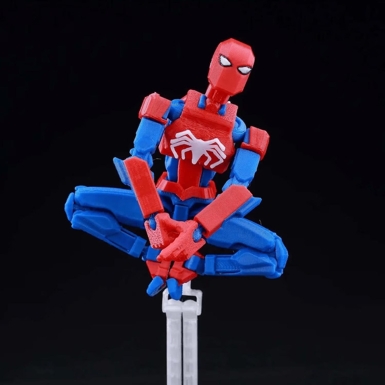 May include: A posable action figure of a character with a red and blue suit, featuring a white spider emblem on the chest. The figure is depicted in a seated, cross-legged position, supported by a clear plastic stand against a black background.