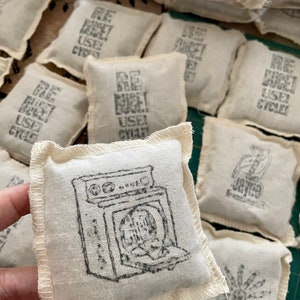 May include: A collection of small, white fabric pouches with various black printed designs. Some pouches feature the text "RE-BAG IT! USE CYCLED", while others have a drawing of a washing machine, a flower, or a bird.