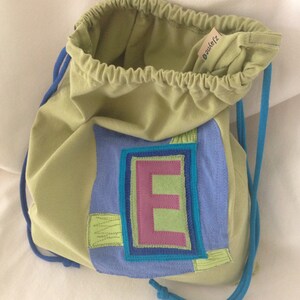May include: A green drawstring bag with a blue and purple patch featuring the letter "E" in a green and pink font. The bag has a blue drawstring.