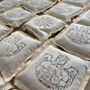 May include: Close-up of several small, square, off-white fabric sachets. Each sachet features a different black ink illustration, including a flower, an angel with a bicycle, and other designs. The edges are finished with a decorative stitch.