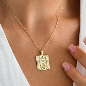 May include: A gold-coloured necklace with a square pendant featuring the letter "R". The pendant has a textured border and hangs from a delicate chain. The necklace is displayed on a person wearing a white top.