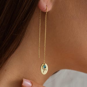 May include: Gold-colored threader earrings. Each earring features a delicate chain that hangs down, with a small, oval pendant at the end. The pendant has a floral design with a teal-colored stone in the center. The earrings are shown on a person.