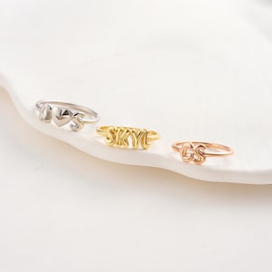 May include: Three rings with personalized designs. One silver ring features the letters "A", a heart, and "S". A gold ring spells out "SKYE". A rose gold ring displays the letters "GS". All rings are on a white surface.