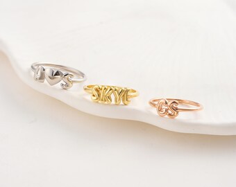 May include: Three rings with personalized designs. One silver ring features the letters "A", a heart, and "S". A gold ring spells out "SKYE". A rose gold ring displays the letters "GS". All rings are on a white surface.
