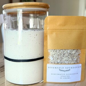 May include: A glass jar with a bamboo lid filled with a white sourdough starter. Next to it is a brown paper pouch with a white label that reads "Sevenfold Sourdough Dehydrated Sourdough Starter 20 Grams".