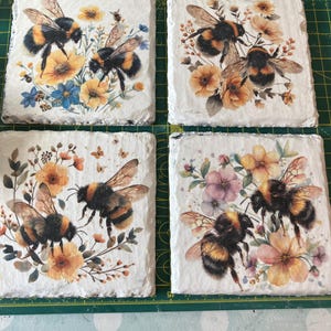 May include: Four square coasters with a white background, each depicting bumblebees and floral arrangements in yellow, orange, blue, and pink. The coasters have a textured, natural appearance, perfect for a coffee table.